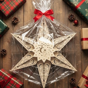 May include: A large, intricately carved wooden star, wrapped in clear cellophane and tied with a red bow. The star features celestial designs and is surrounded by wrapped gifts, pinecones, and ornaments on a wooden surface.