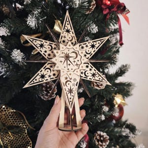 Könnte beinhalten: Ein sternförmiger Weihnachtsbaumaufsatz aus Holz mit aufwendigen Ausschnitten von Sternen, Monden und Halbmonden. Der Stern ist hellbraun mit dunkelbraunen Akzenten und wird vor einem geschmückten Weihnachtsbaum gehalten.