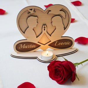 May include: A wooden heart-shaped candle holder with the names "Martin" and "Leonie" engraved on it. The design features silhouettes of a couple holding hands, with a small candle lit in the center. A red rose and rose petals are scattered around.