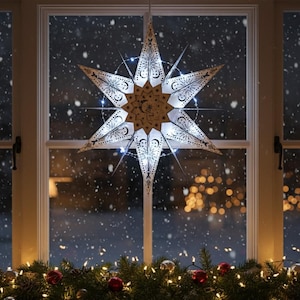 May include: A decorative, illuminated star-shaped window ornament. The star is white with intricate cut-out designs and a central moon and star motif. It is lit with small white lights, creating a warm glow. The star hangs in front of a window with a snowy background.