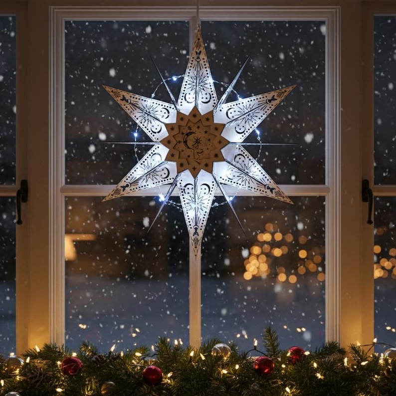 May include: A decorative, illuminated star-shaped window ornament. The star is white with intricate cut-out designs and a central moon and star motif. It is lit with small white lights, creating a warm glow. The star hangs in front of a window with a snowy background.