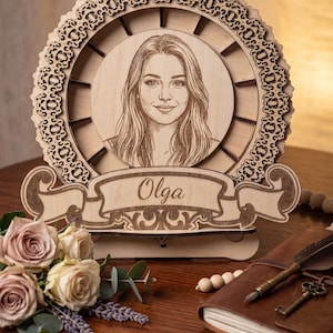 May include: A circular wooden laser-cut frame featuring an intricate lace-like border and a central engraved portrait of a person. Below the portrait, a wooden banner is engraved with the name Olga in a cursive script font, all on a stand.