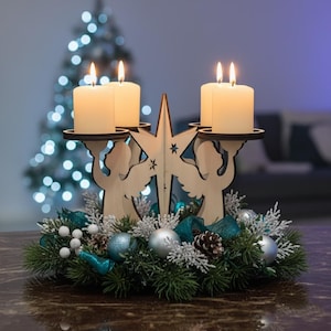 Può includere: Un portacandele natalizio in legno con quattro candele accese. Il supporto presenta sagome di angeli e un design a stella. È decorato con pino, ornamenti e nastro verde acqua, creando un centrotavola festivo.