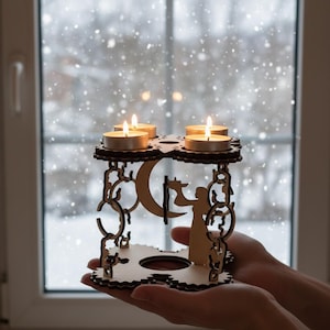 Könnte beinhalten: Ein hölzerner Kerzenhalter mit vier brennenden Teelichtern. Der Halter hat ein dekoratives Design mit einem Mond und einer Personensilhouette. Der Halter wird vor einem Fenster mit fallendem Schnee gehalten.
