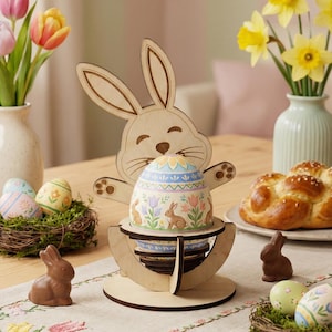 Könnte beinhalten: Ein hölzerner Osterhasen-Eierhalter mit einem verzierten Ei mit Hasen und floralen Mustern. Die Szene umfasst Schokoladenhasen, dekorierte Eier in Nestern und Vasen mit Blumen, wodurch eine festliche Osterdekoration entsteht.