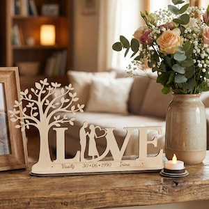 May include: A wooden decorative piece spelling "LOVE", with a tree and a couple silhouette. The base has names and a date. A vase of flowers and a candle are also visible. The piece is made of light-colored wood.