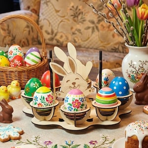 Könnte beinhalten: Ein hölzerner Ostereierhalter mit Hasenmotiv, der dekorierte Eier hält. Ein Korb mit Eiern und Schokoladenhasen ist ebenfalls vorhanden. Die Eier sind mit verschiedenen Mustern und Farben bemalt, darunter Blumenmuster und Streifen.