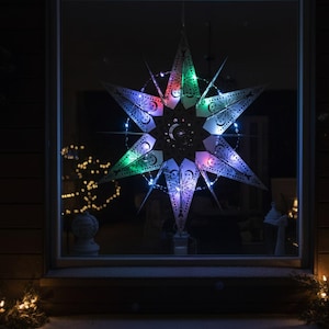 May include: A large, decorative star-shaped light fixture with intricate cut-out patterns and a crescent moon design. The star is illuminated with colorful LED lights in green, red, and purple, creating a festive glow. The star is hanging in a window.