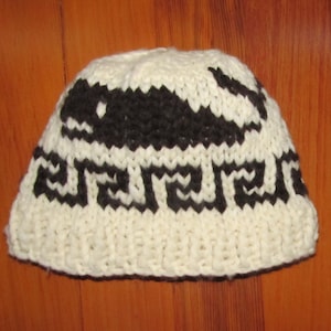 May include: White knitted hat with a black and white geometric design. The design features a stylized whale or fish.