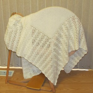 Peut inclure: Châle crème en tricot ajouré, drapé sur un cadre en bois. Le châle présente un motif de dentelle géométrique et un centre carré uni. Les bords ont un design festonné, ajoutant une touche délicate à l'accessoire tricoté.