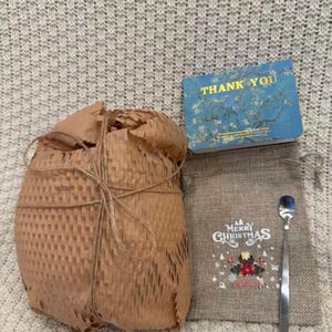 May include: A gift set with a wrapped package, a small burlap bag with "Merry Christmas" text and a spoon, and a blue "Thank You" card with a floral design. The wrapped package is tied with twine.