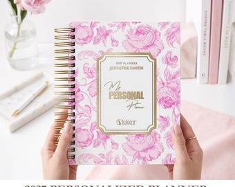 Könnte beinhalten: Ein spiralgebundener Planer für 2027 mit weißem Einband und rosa Blumenmuster. In der Mitte befindet sich ein gold umrandetes rechteckiges Etikett mit dem Text 2027 PLANNER, JENNIFER SMITH, MY PERSONAL Planner und dem Markenlogo lolamir.