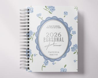 Könnte beinhalten: Ein personalisierter 2026-Planer mit floralem Design. Das weiße Cover zeigt blaue Blumenakzente und ein silbernes Oval mit dem Text "2026 Personal Planner" und dem Namen "Vanessa Smith". Der Planer ist spiralgebunden.
