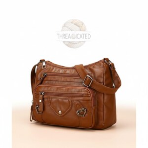 May include: A brown leather crossbody bag with multiple zippered compartments and a heart-shaped accent. The bag has an adjustable shoulder strap and the text "THREADICATED" is visible.