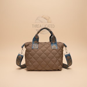 Peut inclure: Sac fourre-tout matelassé marron avec un motif géométrique. Le sac est doté d'une bandoulière et de poignées à motifs bleus et dorés. Le texte "THREADICATED" est au-dessus du sac.