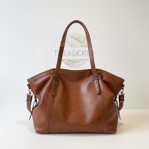 May include: A brown leather tote bag with two top handles and a detachable shoulder strap. The bag has a rectangular shape with a wide opening and is decorated with metal rivets and hardware. The text "TEA OCATED" is visible on the top of the bag.