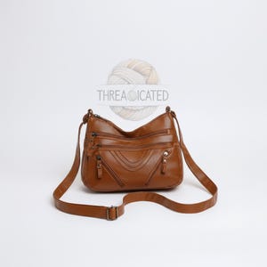 Puede incluir: Un bolso bandolera de cuero marrón con múltiples compartimentos con cremallera y una correa ajustable para el hombro. El bolso tiene un aspecto texturizado y un cierre de hebilla. El texto "THREADICATED" es visible en la parte superior.