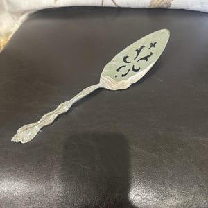 May include: A silver-toned metal cake server featuring an elongated, triangular blade with a decorative cut-out pattern. The handle has a scalloped, embossed design with intricate floral-like detailing at the base and along the stem.