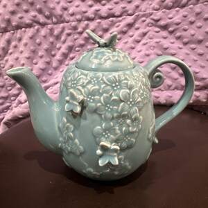 Puede incluir: Una tetera de cerámica azul claro con un patrón floral en relieve de flores de cerezo y pequeñas mariposas en el cuerpo. La tapa está rematada con un pequeño asa en forma de mariposa, y la tetera cuenta con un pico y un asa curvados.