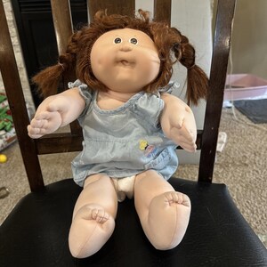 May include: A doll with a round face, dimpled cheeks, and brown yarn hair styled in two pigtails. The doll has a light skin tone and is wearing a light blue sleeveless dress with a small embroidered yellow and pink flower detail on the front.