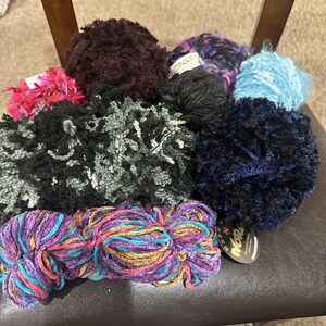 May include: A collection of assorted yarn balls in various textures and colors, including dark brown, black, light blue, and a multi-colored skein with purple, pink, and gold tones. Some balls feature labels with text such as Noro and Ironstone.