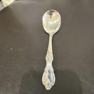 Puede incluir: Una cuchara de metal plateado pulido con un patrón floral y de volutas en relieve en el mango. La cuchara tiene un cuenco redondeado y un cuello estrecho, con intrincados detalles decorativos que se extienden hasta la base del mango.