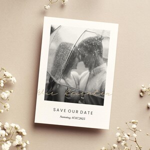 May include: A black and white photo of a couple under an umbrella. The text "Save Our Date" is printed in black and the date "Samstag 07.07.2025" is printed in black below the text.