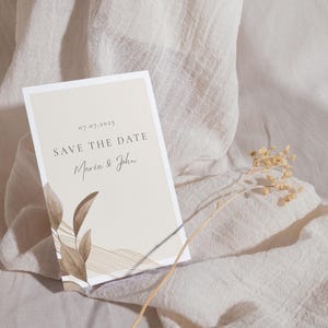 May include: A beige save the date card with brown calligraphy that reads "Save the Date" and "Maria & John" with the date "07.07.2025". The card has a brown leaf design and a thin brown line pattern.
