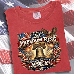May include: A red t-shirt featuring a graphic print of the Liberty Bell, a bald eagle, and American flags. The text reads Let Freedom Ring and Celebrating 250 Years of American Independence. The shirt has a Comfort Colors brand label at the neck.