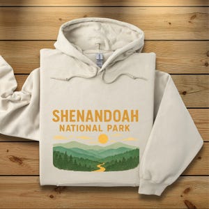 A folded cream-colored hoodie displays a yellow and green Shenandoah National Park graphic centered on the chest.