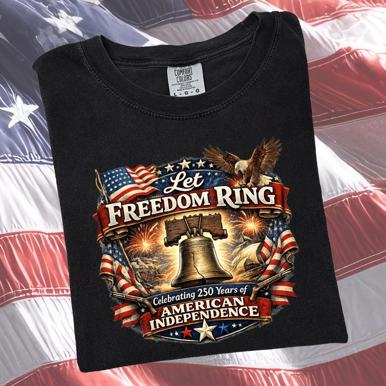 May include: A black t-shirt featuring a graphic print of the Liberty Bell, a bald eagle, and American flags. The text reads Let Freedom Ring and Celebrating 250 Years of American Independence. The shirt has a Comfort Colors brand label at the neck.