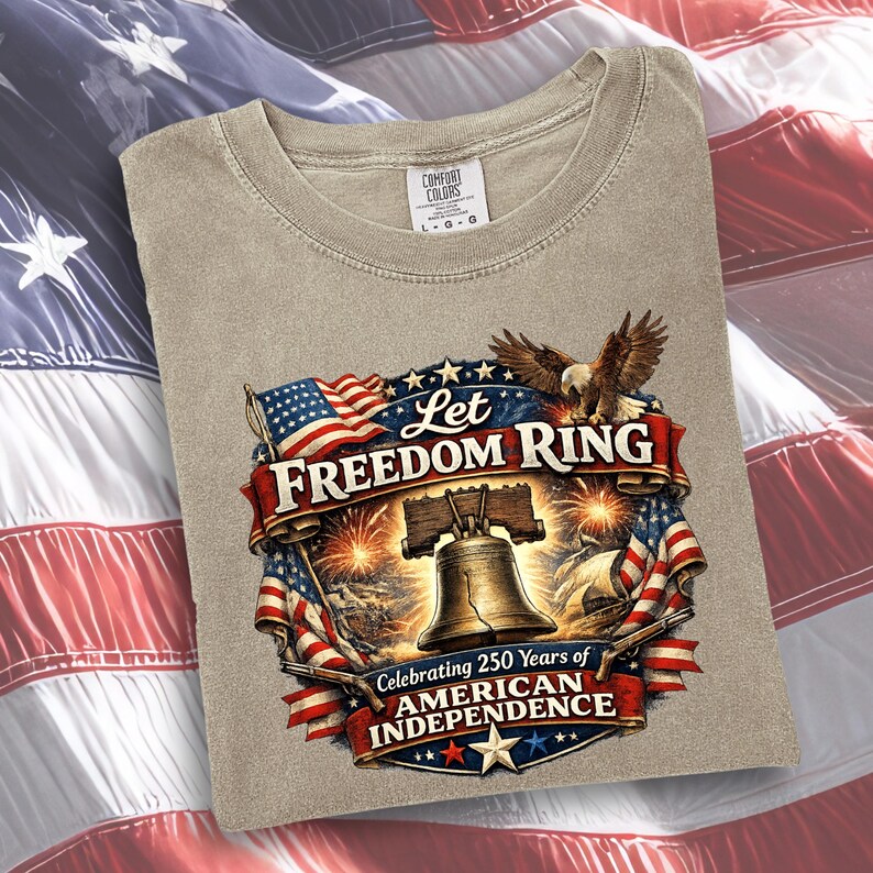 May include: A beige t-shirt featuring a graphic print of the Liberty Bell, a bald eagle, and American flags. The text reads Let Freedom Ring and Celebrating 250 Years of American Independence. The shirt has a Comfort Colors brand neck label.