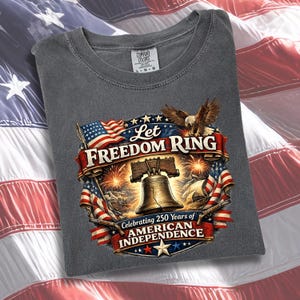 May include: A charcoal grey t-shirt featuring a graphic print of the Liberty Bell, a bald eagle, and American flags. The text reads Let Freedom Ring and Celebrating 250 Years of American Independence. The design includes fireworks and a star.