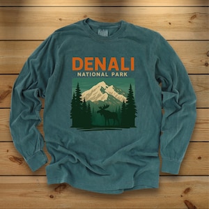 Pode incluir: Uma camisola de manga comprida em verde-azulado com uma impressão gráfica que inclui o texto DENALI NATIONAL PARK em laranja. O gráfico mostra um pico nevado, pinheiros verde-escuros e a silhueta de um alce em primeiro plano.