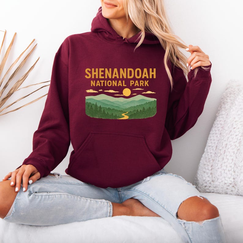 A person wearing a dark maroon hoodie with a Shenandoah National Park graphic, sitting cross-legged on a bed.