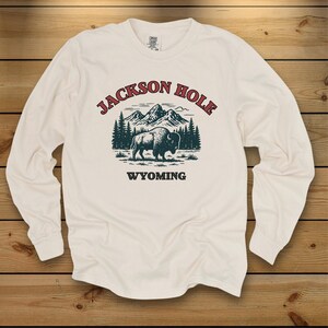 May include: A cream-colored long-sleeve shirt featuring a graphic print of a bison standing in front of a mountain range and pine trees. The text "JACKSON HOLE" is arched above the illustration in red, and "WYOMING" is printed in black below.