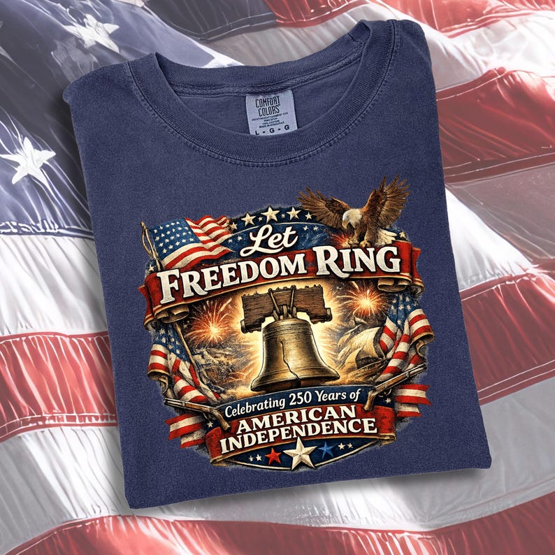 May include: A navy blue t-shirt featuring a graphic print of the Liberty Bell, a bald eagle, and American flags. The text reads Let Freedom Ring and Celebrating 250 Years of American Independence. The shirt has a Comfort Colors brand label.