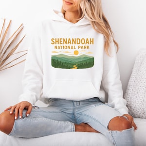 A person wearing a white hooded sweatshirt with a Shenandoah National Park graphic, sitting on a white surface.