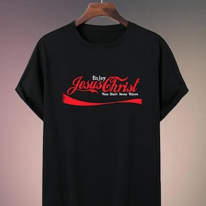 May include: Black t-shirt with the words "Enjoy Jesus Christ" in red script, with "Thou Shalt Never Thirst" below. The design is reminiscent of a popular beverage logo. The shirt is hanging on a wooden hanger.