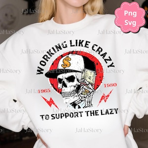 May include: White sweatshirt with a graphic featuring a skull wearing a backwards baseball cap and holding money. The text reads "WORKING LIKE CRAZY TO SUPPORT THE LAZY" with the years 1965 and 1980.