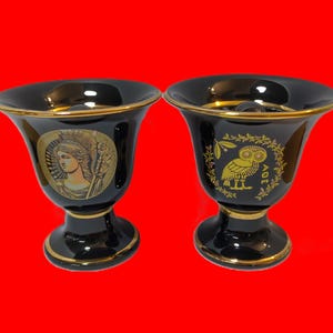 May include: Two black ceramic decorative cups with gold trim. One cup features a gold image of a woman, the other an owl. The cups have a flared top and a pedestal base. The background is red.
