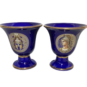 Puede incluir: Dos jarrones de cerámica azul y dorado con diseños de inspiración griega. Un jarrón presenta un casco dorado y el otro un perfil dorado de una mujer.