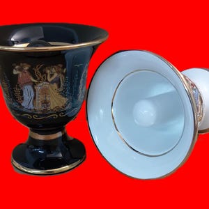Pode incluir: Dois vasos de cerâmica preto e branco com detalhes dourados. O vaso à esquerda tem uma cena de três mulheres pintadas nele. O vaso à direita é branco com uma borda dourada.