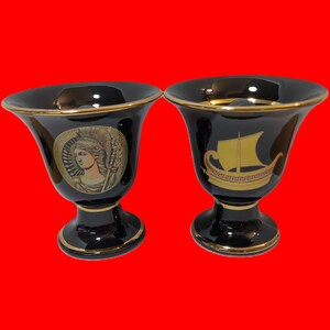 May include: Two black ceramic cups with gold trim and painted designs. One cup features a woman with a helmet and a spear, the other features a gold ship with red accents.