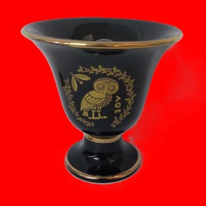 Könnte beinhalten: Eine schwarze Keramikvase mit goldfarbenem Rand und einem goldenen Eulenmotiv. Die Eule ist von einem Kranz aus Olivenzweigen umgeben und das Wort "AOE" steht darunter.