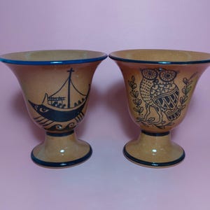 Könnte beinhalten: Zwei Keramikbecher mit blauen und braunen Designs. Ein Becher zeigt ein Schiff mit Menschen an Bord und der andere Becher zeigt eine Eule.
