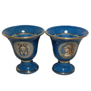 May include: Two matching teal-colored ceramic vases with gold trim. Each vase features a classical design with a gold and white illustration of a helmet and a woman's face. The vases have a flared top and a pedestal base.