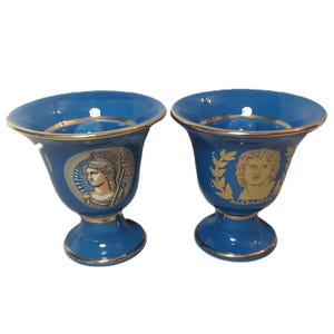 Puede incluir: Dos tazas de cerámica azul con ribetes dorados e imágenes de figuras griegas. La taza de la izquierda tiene una imagen de una mujer con casco y una lanza. La taza de la derecha tiene una imagen de un hombre con una corona de laurel y la palabra "ALEXANDER".