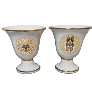 May include: Two white ceramic vases with gold trim. One vase features a Medusa head design and the other features a golden helmet design.