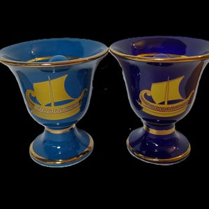 May include: Two ceramic cups with gold trim and a gold ship design. One cup is blue and the other is dark blue.